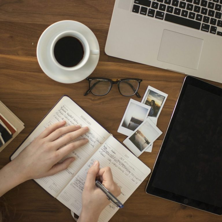 Main en train d'écrire dans un carnet, café et ordinateur portable à proximité.