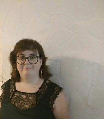 Femme aux cheveux bruns, portant des lunettes et un haut noir, devant un mur clair.