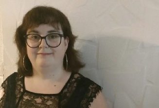 Femme avec des lunettes, cheveux bruns ondulés, portant une blouse noire à dentelle.